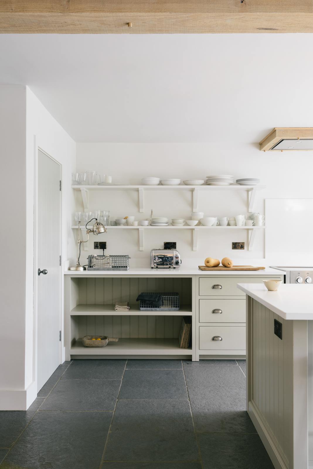 A quiet corner of a tranquil and calm Shaker kitchen features a long sink and cooker run with a Silestone worktop and splashback, a long kitchen island with a breakfast nook, and shelves. Beautifully decorated with muted ceramics, a wooden chopping board, butternut squashes,  and a silver toaster, this kitchen creates a lovely, simple space that embraces natural materials.
