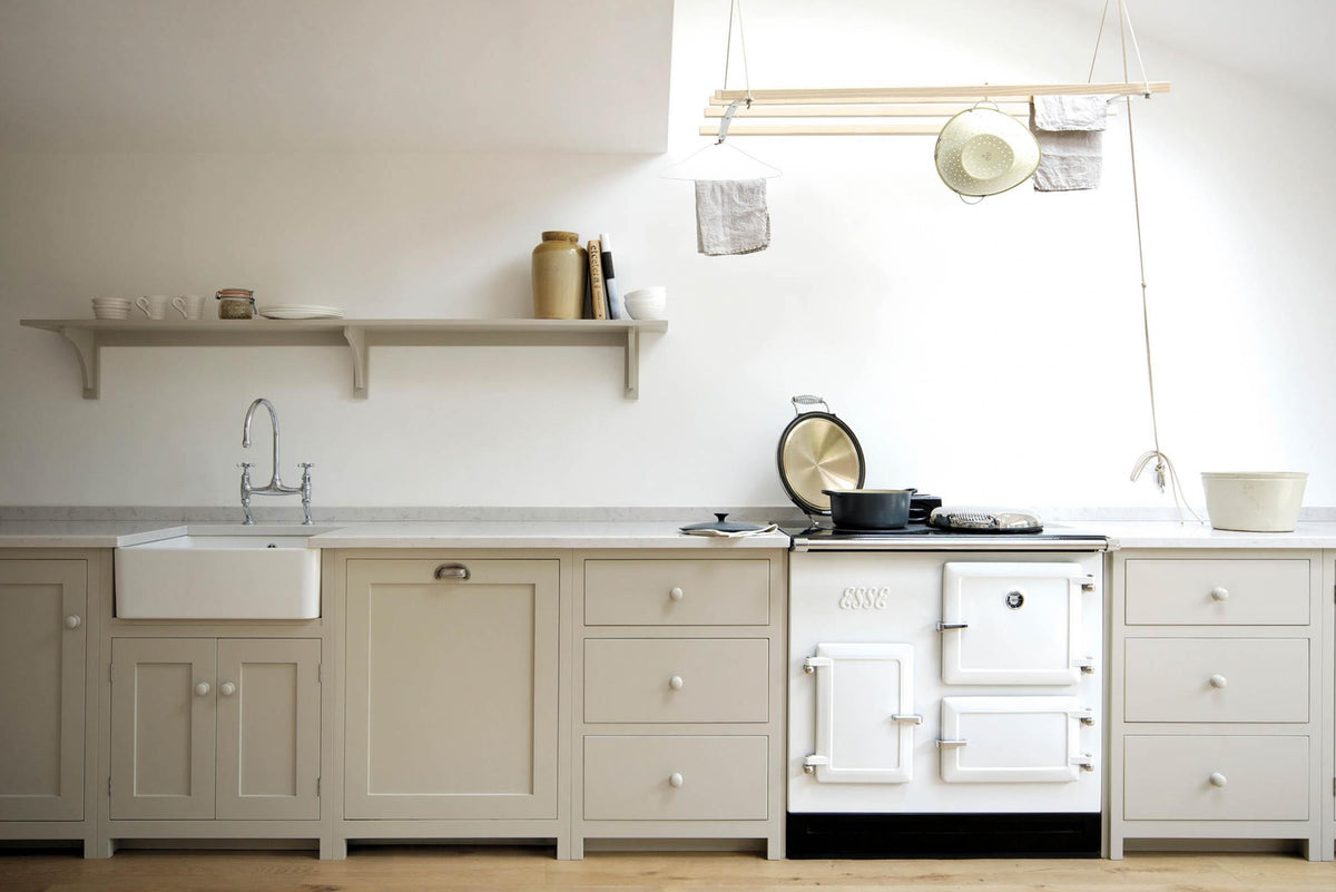 An elegant mushroom-coloured Shaker kitchen features a sink and cooker run with a Carrara marble worktop, a white gas cooker, a single Belfast sink with an antique silver Ionian tap, and a shelf. Adorned with cast iron pots, artisan ceramics, simple plates and mugs, and a deVOL laundry maid, it creates a calm and functional space.