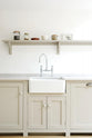 An elegant mushroom-coloured Shaker kitchen features a sink and cooker run with a Carrara marble worktop, a single Belfast sink with an antique silver Ionian tap, and a shelf. Adorned with dried ingredients in a clean jar, artisan ceramics and a small framed picture of olives, it creates a calm and functional space.