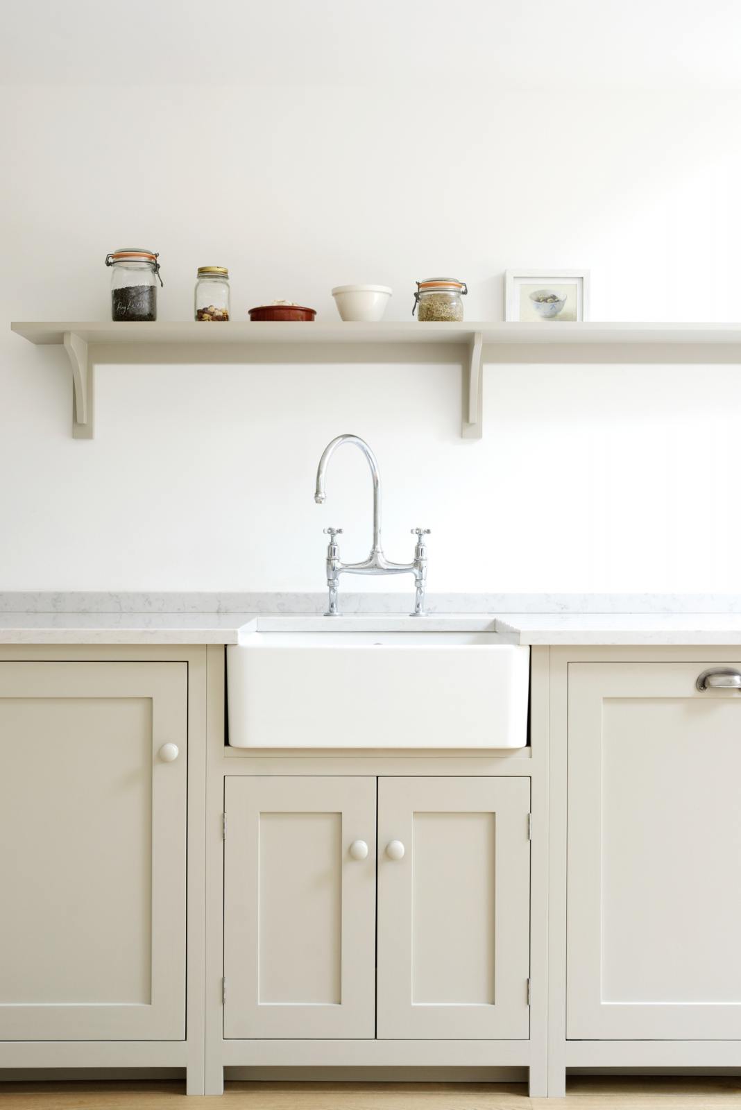 An elegant mushroom-coloured Shaker kitchen features a sink and cooker run with a Carrara marble worktop, a single Belfast sink with an antique silver Ionian tap, and a shelf. Adorned with dried ingredients in a clean jar, artisan ceramics and a small framed picture of olives, it creates a calm and functional space.