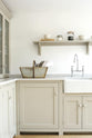 A corner of an elegant mushroom-coloured Shaker kitchen features a sink and cooker run with a Carrara marble worktop, a single Belfast sink with an antique silver Ionian tap, a shelf, and glazed countertop cupboards. Adorned with dried ingredients in a clear jar, artisan ceramics, and a vintage wooden basket with towels inside, it creates a calm and functional space.