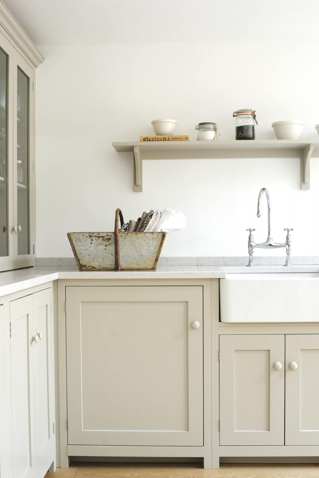 A corner of an elegant mushroom-coloured Shaker kitchen features a sink and cooker run with a Carrara marble worktop, a single Belfast sink with an antique silver Ionian tap, a shelf, and glazed countertop cupboards. Adorned with dried ingredients in a clear jar, artisan ceramics, and a vintage wooden basket with towels inside, it creates a calm and functional space.