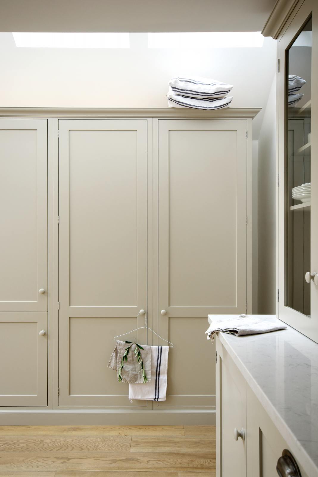 A corner of an elegant mushroom-coloured Shaker kitchen features a sink and cooker run with glazed countertop cupboards, and a bank of full-height cupboards. Adorned with simple plates, tea towels, pillows and pillow cases, it creates a calm and functional space.