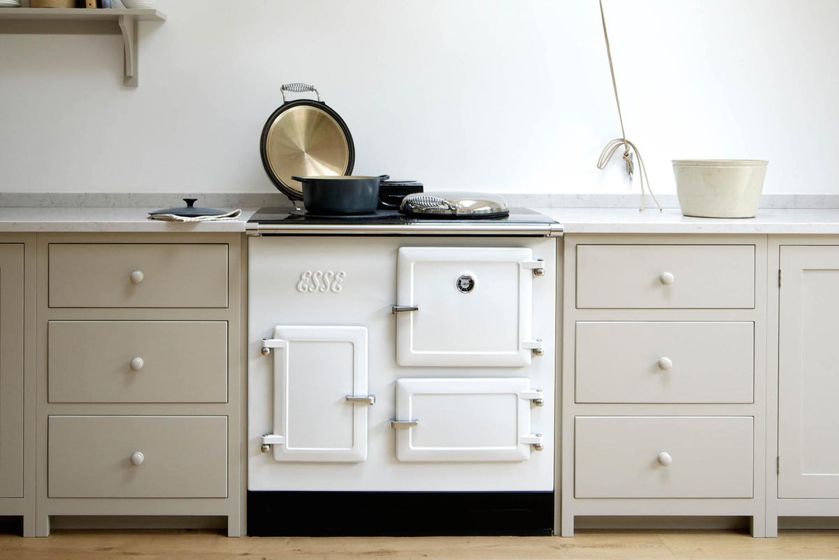 An elegant mushroom-coloured Shaker kitchen features a sink and cooker run with a Carrara marble worktop, a white gas cooker, and a shelf. Adorned with cast iron pots, artisan ceramics, and a deVOL laundry maid, it creates a calm and functional space.
