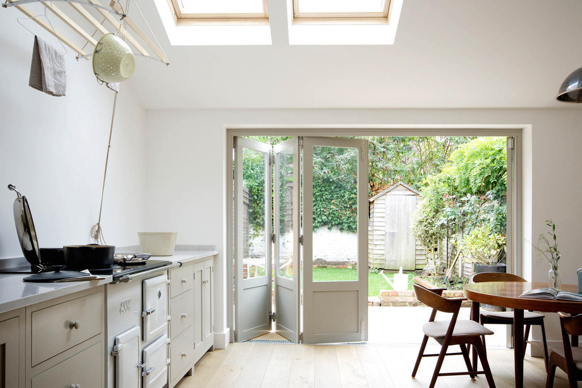 An elegant mushroom-coloured Shaker kitchen features a sink and cooker run with a Carrara marble worktop, a white gas cooker, a rustic wooden dining table and chairs, and bifold patio doors leading to the luscious garden. Adorned with cast iron pots, artisan ceramics, and a deVOL laundry maid, it creates a calm and functional space.