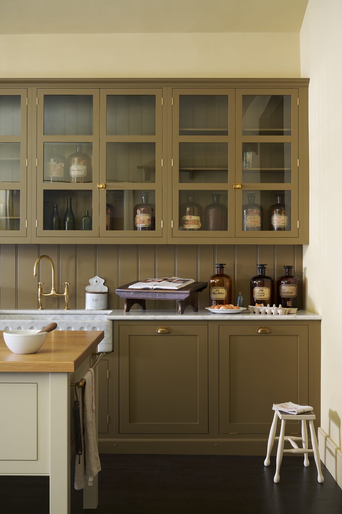A quiet corner of a simple Quaker cream and Folgate green Shaker kitchen features a cooker and sink run with Carrara marble worktop, glazed wall cupboards, and a bespoke prep table with a classic oak worktop. Lovingly adorned with milk pendant lights, artisan ceramics, a fresh carton of eggs, and antique furniture, it creates a warm and inviting atmosphere.