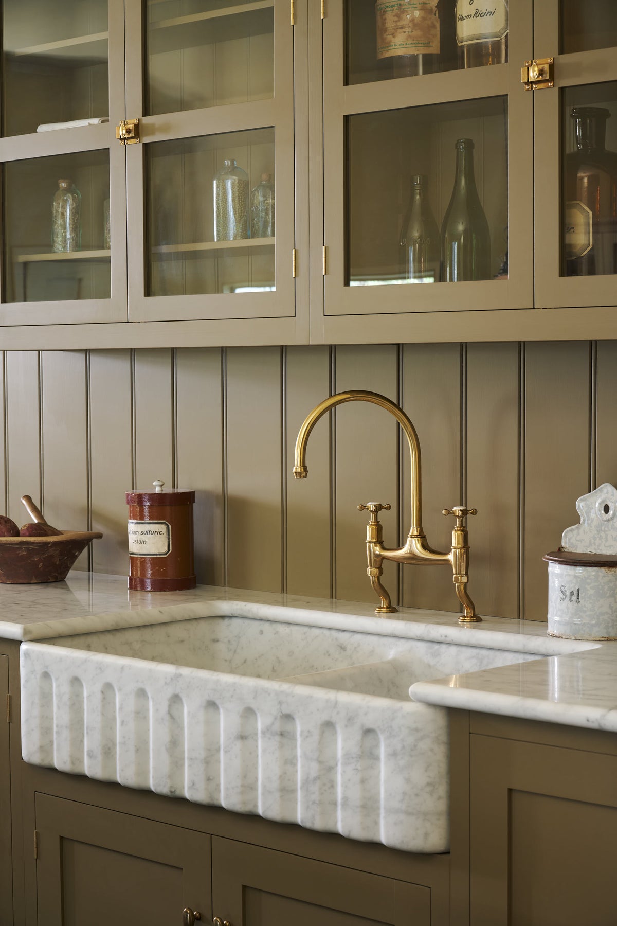 A close-up and oblique view of a simple Quaker cream and Folgate green Shaker kitchen features a cooker and sink run with Carrara marble worktop, an aged brass Ionian tap, and glazed wall cupboards. Lovingly adorned with artisan ceramics, and antiques, it creates a warm and inviting atmosphere.
