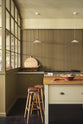 A quiet corner of a simple Quaker cream and Folgate green Shaker kitchen features a cooker and sink run with Carrara marble worktop, and a bespoke prep table with a classic oak worktop. Lovingly adorned with milk pendant lights, artisan ceramics, fresh potatoes, Helen stools, and antiques, it creates a warm and inviting atmosphere.
