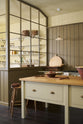 An oblique view of a quiet corner of a simple Quaker cream and Folgate green Shaker kitchen features a cooker and sink run with Carrara marble worktop, and a bespoke prep table with a classic oak worktop. Lovingly adorned with milk pendant lights, artisan ceramics, fresh potatoes, Helen stools, and antiques, it creates a warm and inviting atmosphere.