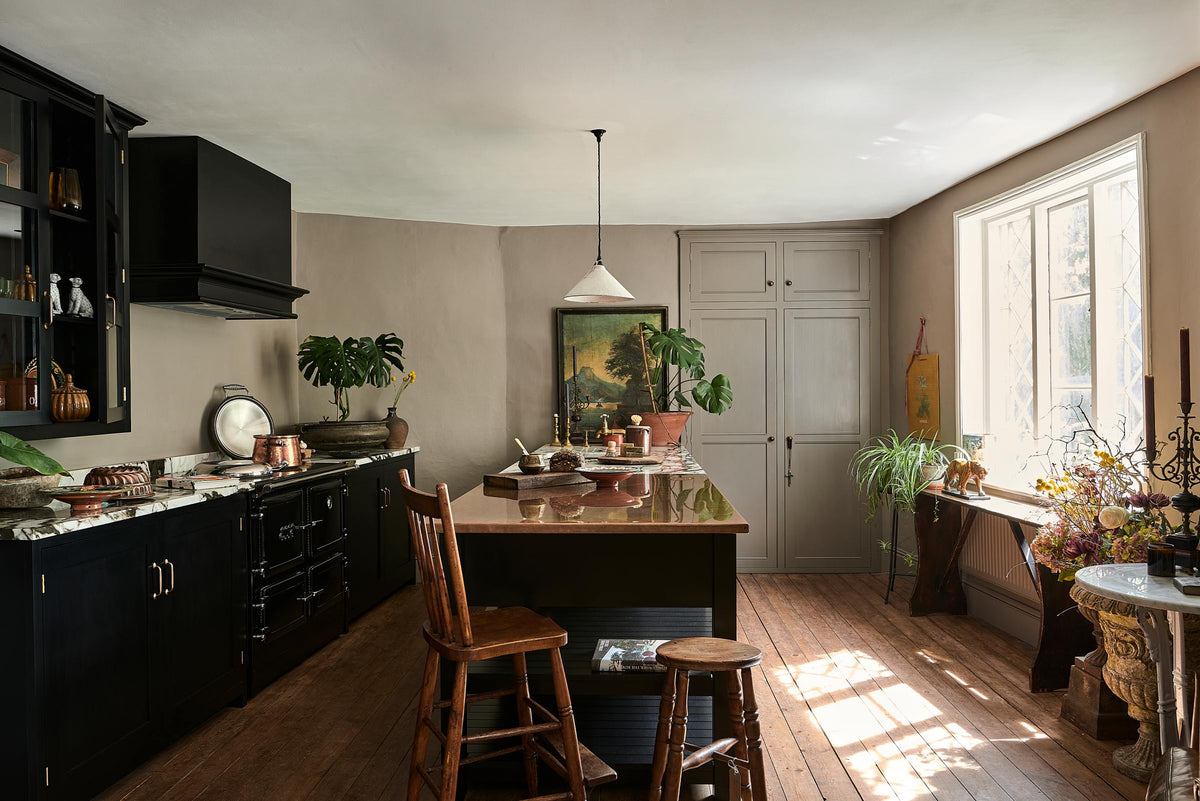 Bright light floods an understated Painter’s Black Shaker kitchen featuring an aged copper sink, aged brass taps, and a San Simone quartzite worktop—its pale white base accented with bold dark grey veins and subtle gold flecks. Grey walls frame the space, with a cheese plant and vintage oil painting adding character in the background.