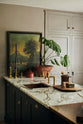 A closer look at a painter’s black shaker kitchen’s sink run with an aged copper sink, aged brass taps and a San Simone quartzite worktop - this stone has a pale white base with bold dark grey veining and little flickers of gold throughout. The walls are painted grey, and there is a cheese plant and a vintage oil painting in the background.