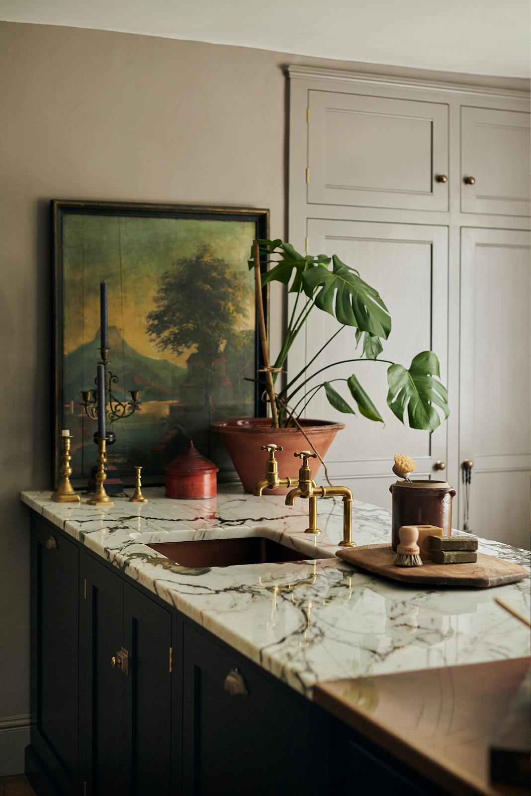 A closer look at a painter’s black shaker kitchen’s sink run with an aged copper sink, aged brass taps and a San Simone quartzite worktop - this stone has a pale white base with bold dark grey veining and little flickers of gold throughout. The walls are painted grey, and there is a cheese plant and a vintage oil painting in the background.