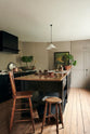 Bright light pours into an understated painter’s black shaker kitchen featuring an aged copper sink, aged brass taps, and a San Simone quartzite worktop—its pale white base accented with bold dark grey veins and subtle gold flecks. Grey walls frame the space, with a cheese plant and vintage oil painting adding character in the background.
