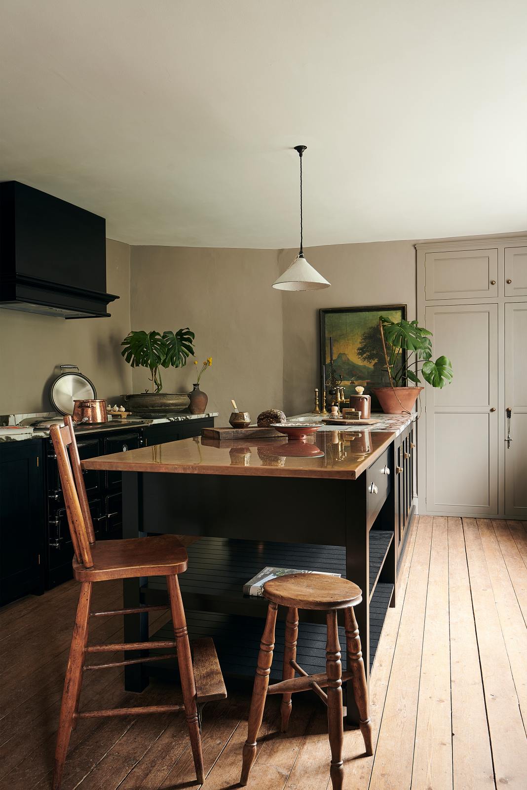 Bright light pours into an understated painter’s black shaker kitchen featuring an aged copper sink, aged brass taps, and a San Simone quartzite worktop—its pale white base accented with bold dark grey veins and subtle gold flecks. Grey walls frame the space, with a cheese plant and vintage oil painting adding character in the background.