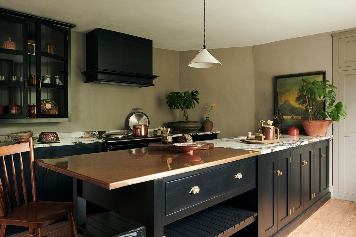 An oblique view of an understated painter’s black shaker kitchen featuring an aged copper sink, aged brass taps, and a San Simone quartzite worktop—its pale white base accented with bold dark grey veins and subtle gold flecks. Grey walls frame the space, with a cheese plant and vintage oil painting adding character in the background.