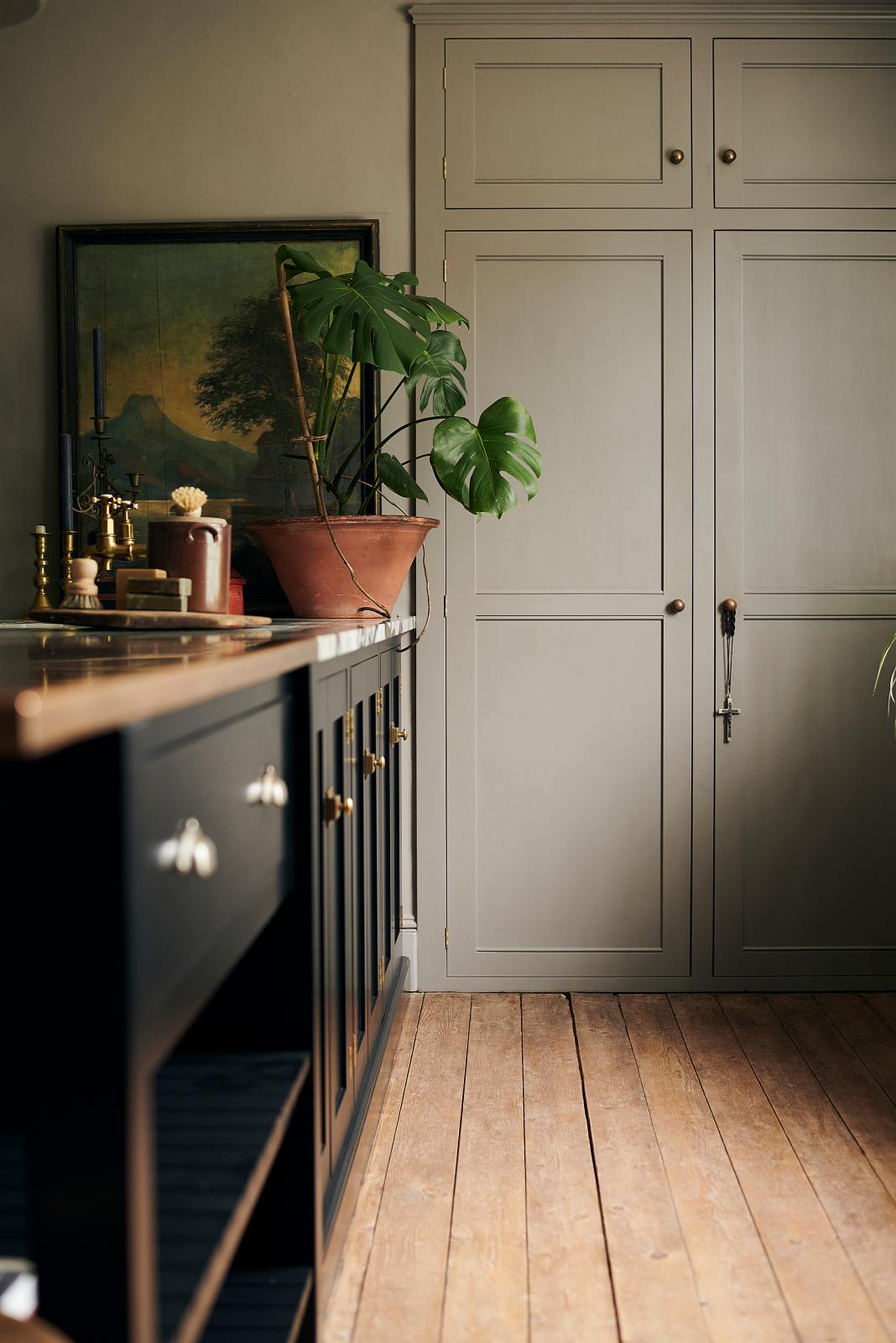 Bright light pours into an understated painter’s black shaker kitchen with a vintage built-in cabinet, a painting on the worktop, a cheese plant, and elegant candles.