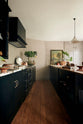 A straight view of an understated painter’s black shaker kitchen featuring an aged copper sink, aged brass taps, and a San Simone quartzite worktop—its pale white base accented with bold dark grey veins and subtle gold flecks. Grey walls frame the space, with a cheese plant and vintage oil painting adding character in the background.
