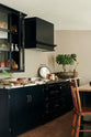 An oblique view of an understated painter’s black shaker kitchen featuring an aged copper sink, aged brass taps, and a San Simone quartzite worktop—its pale white base accented with bold dark grey veins and subtle gold flecks. Grey walls frame the space, with a cheese plant and vintage oil painting adding character in the background.