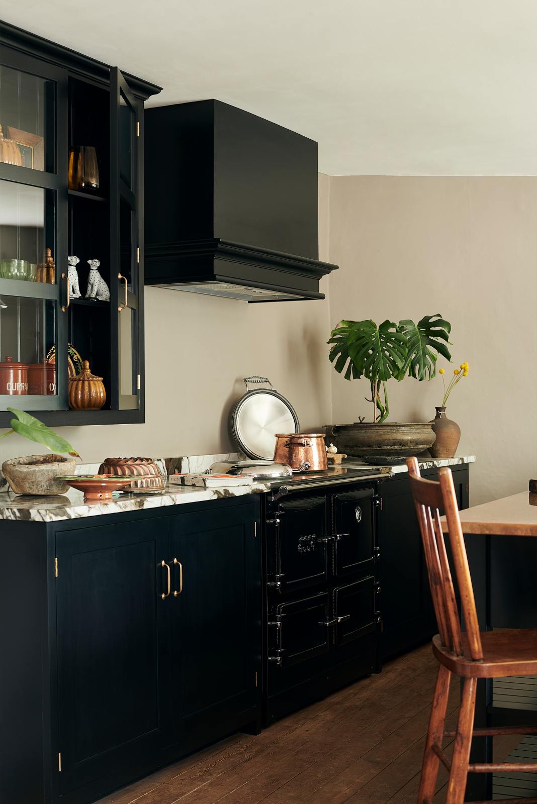 An oblique view of an understated painter’s black shaker kitchen featuring an aged copper sink, aged brass taps, and a San Simone quartzite worktop—its pale white base accented with bold dark grey veins and subtle gold flecks. Grey walls frame the space, with a cheese plant and vintage oil painting adding character in the background.