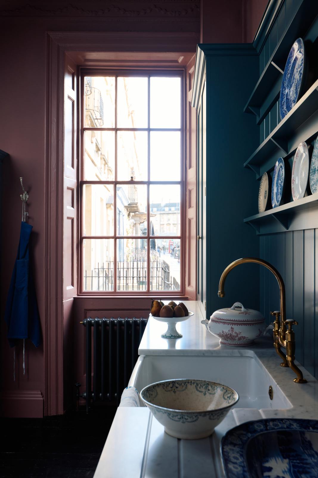 Looking across the sink run and through the adjacent sash window, a glimpse of Bath’s classic Georgian architecture unfolds outside. Light floods the room, casting a soft sheen on the painted teal cabinetry and Carrara marble worktop. The deep gold aged brass tap catches the light, adding dramatic warmth to the scene.