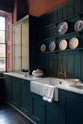 An oblique view of a sink run which shows the furniture, fittings and styling in more detail. The knobs are small, delicate and oval-shaped, and the integrated dishwasher to the left of the sink has a classic cup handle in the centre of the top rail. The space has an almost regal feeling with the moody wine walls, deep blue tones and the rich gold of the aged brass tap and fittings.