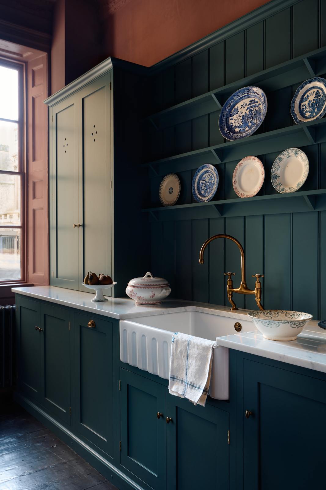 An oblique view of a sink run which shows the furniture, fittings and styling in more detail. The knobs are small, delicate and oval-shaped, and the integrated dishwasher to the left of the sink has a classic cup handle in the centre of the top rail. The space has an almost regal feeling with the moody wine walls, deep blue tones and the rich gold of the aged brass tap and fittings.