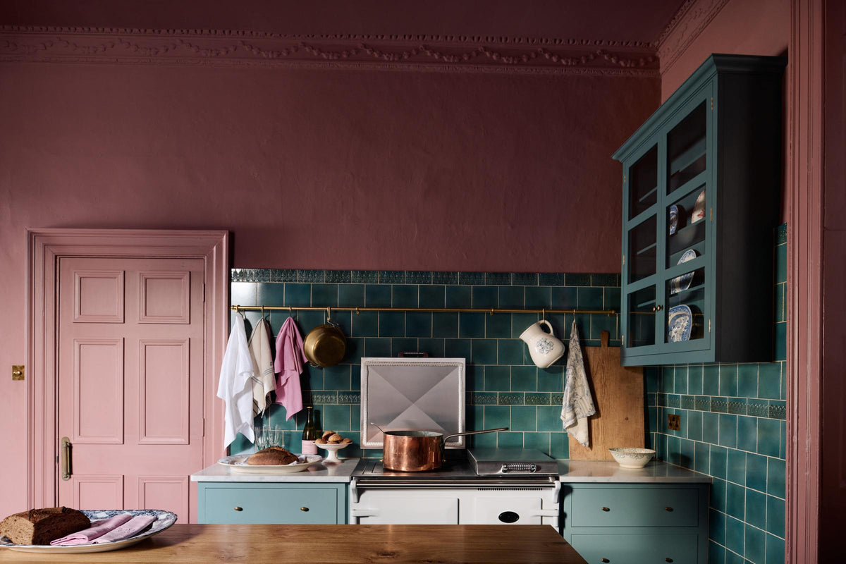 A cooker run that is small and compact with Shaker pan drawers on either side of a white Aga range cooker and a large old copper saucepan on the stove. The wall above is adorned with our glossy vintage teal lace market Tiles, an aged brass hanging rail, and a grand London Glazed Wall Cupboard on the adjacent wall. To the left of the run is a door with beautiful original Georgian moulding and architrave, painted in the same dusky wine colour as the walls.