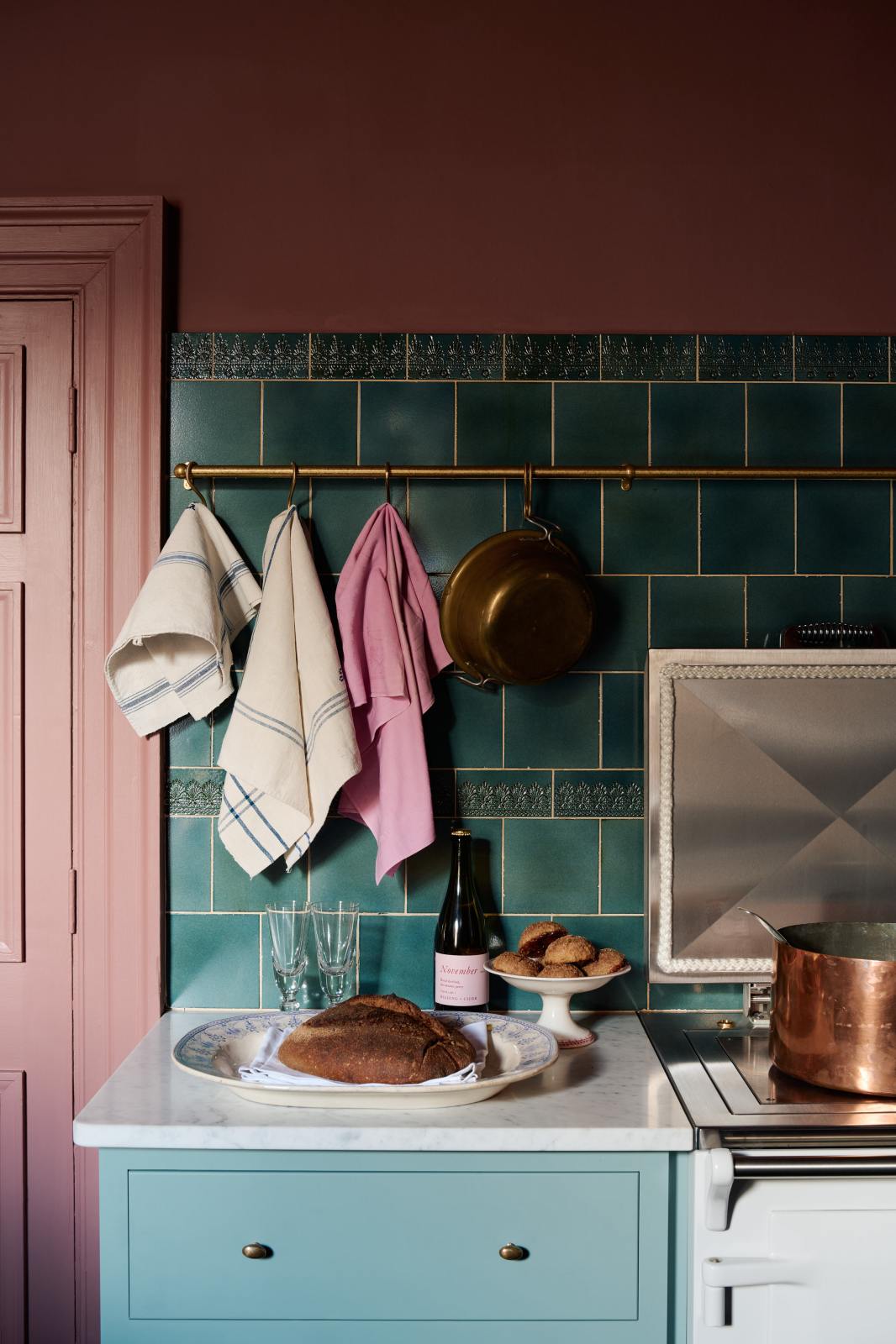 A large display of glossy Lace Market splashback tiles in rich vintage teal contrasts beautifully with wine-coloured walls and aged brass hanging rails adorned with vintage tea towels. In the foreground, a bottle of wine, freshly baked bread, and pastries rest on the Carrara marble worktop of the Clerkenwell Blue Shaker kitchen’s cooker run.