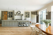 A grand warm-grey Shaker kitchen features a cooker run with a Carrara marble worktop, glazed wall cabinets, an overmantle canopy, and a wide kitchen island with a single sink. Adorned with beloved furniture, simple white platters, glass pendant lights, and pale wood herringbone floors, the space feels sophisticated and cool.