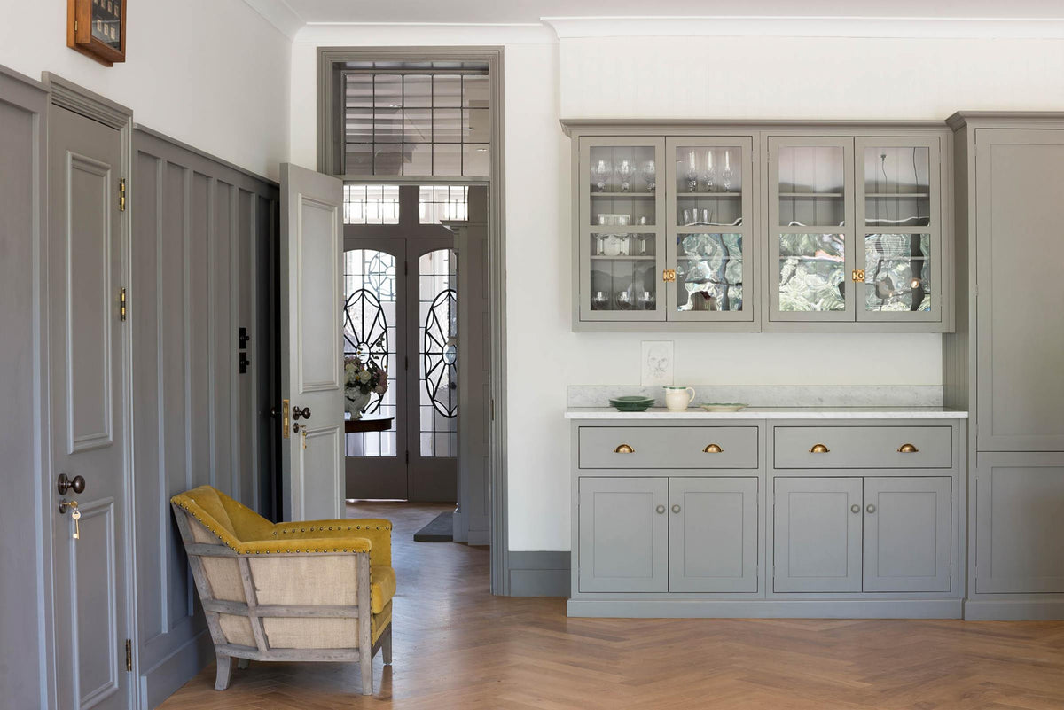 A quiet corner of a grand warm-grey Shaker kitchen features a pantry run with a Carrara marble worktop and glazed wall cabinets. Adorned with beloved furniture, simple white platters, a yellow armchair, and pale wood herringbone floors, the space feels effortlessly sophisticated and cool.