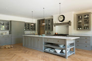 An angled view of a grand warm-grey Shaker kitchen features a cooker run with a Carrara marble worktop, glazed wall cabinets, an overmantle canopy, a wide kitchen island with a single sink, and a pantry run. Adorned with beloved furniture, simple white platters, glass pendant lights, and pale wood herringbone floors, the space feels sophisticated and cool.