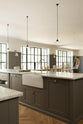 An angled view of a grand warm-grey Shaker kitchen features a cooker run with a Carrara marble worktop, and a wide kitchen island with a single sink,  and a chrome Ionian tap. Adorned with beloved furniture, simple white platters, glass pendant lights, and pale wood herringbone floors, the space feels sophisticated and cool.