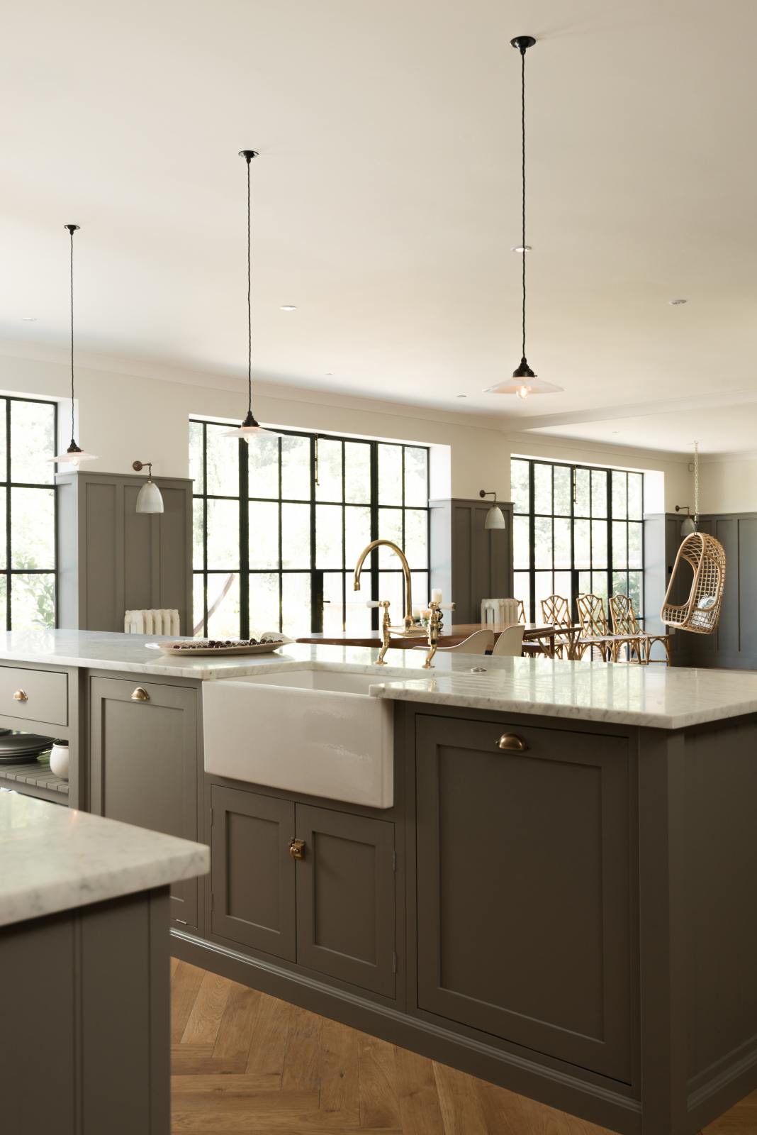 An angled view of a grand warm-grey Shaker kitchen features a cooker run with a Carrara marble worktop, and a wide kitchen island with a single sink,  and a chrome Ionian tap. Adorned with beloved furniture, simple white platters, glass pendant lights, and pale wood herringbone floors, the space feels sophisticated and cool.