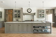 A grand warm-grey Shaker kitchen features a cooker run with a Carrara marble worktop, glazed wall cabinets, an overmantle canopy, and a wide kitchen island with a single sink. Adorned with beloved furniture, simple white platters, glass pendant lights, and pale wood herringbone floors, the space feels sophisticated and cool.