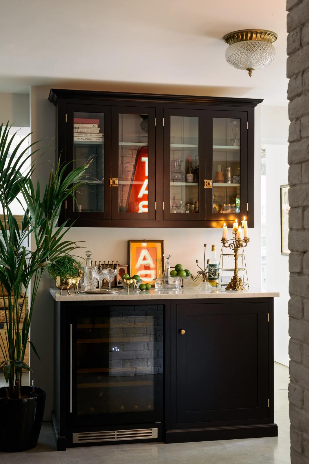 A classy cellar bar features glazed wall cupboards and a small wine cooler run with a Carrara marble worktop. Adorned with golden embellishments such as candelabras, a cake stand, deer ornaments, and a vintage red Tabac neon lights, it creates a sophisticated and social space.