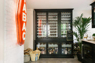 A classy cellar bar features a bank of curiosity cupboards, glazed wall cupboards, and a small wine cooler run with a Carrara marble worktop. Adorned with golden embellishments such as deer ornaments, serving trays, and a vintage red Tabac neon lights, it creates a sophisticated and social space.