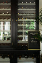 A closer look at a classy cellar bar’s curiosity cupboards reveals a vintage wine collection resting on wine racks. Adorned with a tall house plant and a golden-framed peg letter board, it creates a sophisticated and social space.