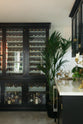 A classy cellar bar features a bank of curiosity cupboards, glazed wall cupboards, and a small wine cooler run with a Carrara marble worktop. Adorned with golden embellishments such as candelabras, a cake stand, deer ornaments, and a golden-framed gentleman's silhouette illustration, it creates a sophisticated and social space.
