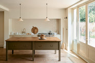 A light, clean, and simple shaker kitchen features a sink and cooker run nestled in a chimney breast, cream porcelain splashback tiles, wooden floorboards, and white walls. Cream cabinets complement an olive green Georgian-style prep table with deep drawers and a rustic oak worktop at the room’s centre, illuminated by two elegant creamware pendant lights.