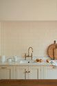 A close-up of the light, clean Shaker kitchen’s single sink with an aged brass Ionian tap set into a marble worktop. The concave wall, lined with cream porcelain splashback tiles, is styled with oranges, wooden chopping boards, ceramic bowls, cups, and a white dish towel.