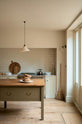 A light, clean, and simple shaker kitchen features a sink and cooker run nestled in a chimney breast, cream porcelain splashback tiles, wooden floorboards, and white walls. Cream cabinets complement an olive green Georgian-style prep table with deep drawers and a rustic oak worktop at the room’s centre, with an elegant creamware pendant light.