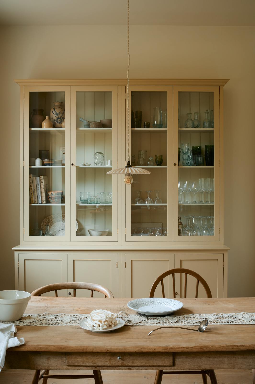 A cream, glazed pantry cupboard stands proudly at the back of a shaker kitchen. In the foreground is an old pine table and antique stick-back chairs, styled with vintage lace, an antique silver spoon and deVOL’s heirloom round platter. A delicate frilly pendant light hangs above the table.