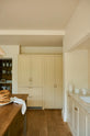 A light, clean, and simple shaker kitchen features a sink and cooker run nestled in a chimney breast, cream porcelain splashback tiles, wooden floorboards, and white walls. Cream full-height cabinets complement an olive green Georgian-style prep table with deep drawers and a rustic oak worktop at the room’s centre, with an elegant creamware pendant light.