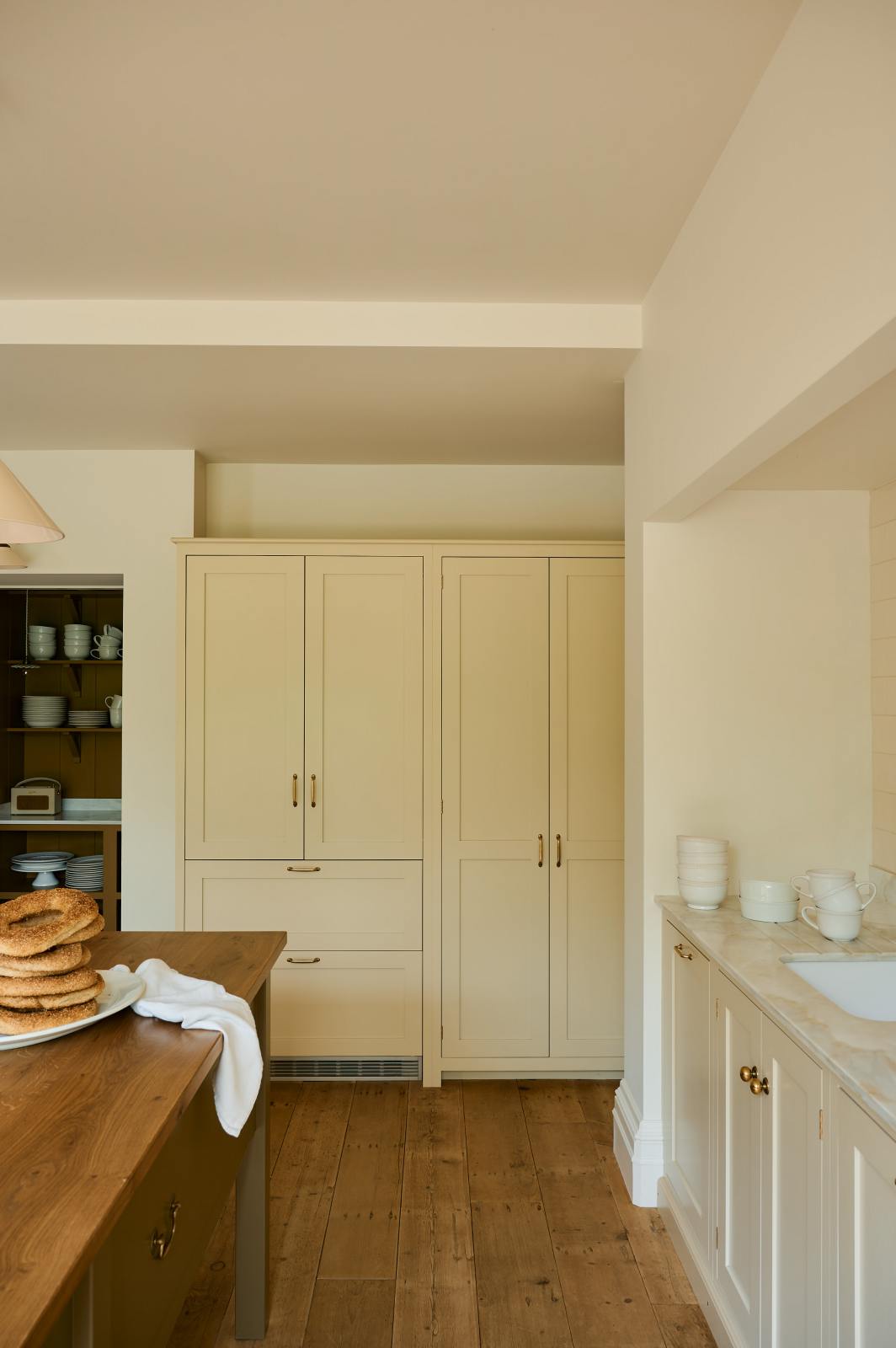 A light, clean, and simple shaker kitchen features a sink and cooker run nestled in a chimney breast, cream porcelain splashback tiles, wooden floorboards, and white walls. Cream full-height cabinets complement an olive green Georgian-style prep table with deep drawers and a rustic oak worktop at the room’s centre, with an elegant creamware pendant light.