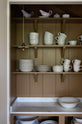 A closer look at a soft brown open pantry cupboard with three top shelves and open cabinets topped with a marble worktop. The space is filled with white ceramic plates, bowls, mugs, a butter dish, and gravy boats. Inside, a small, wide-pleat porcelain light adds a gentle glow.