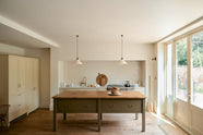 A light, clean, and simple shaker kitchen features a sink and cooker run nestled in a chimney breast, cream porcelain splashback tiles, wooden floorboards, and white walls. Cream cabinets complement an olive green Georgian-style prep table with deep drawers and a rustic oak worktop at the room’s centre, illuminated by two elegant creamware pendant lights.
