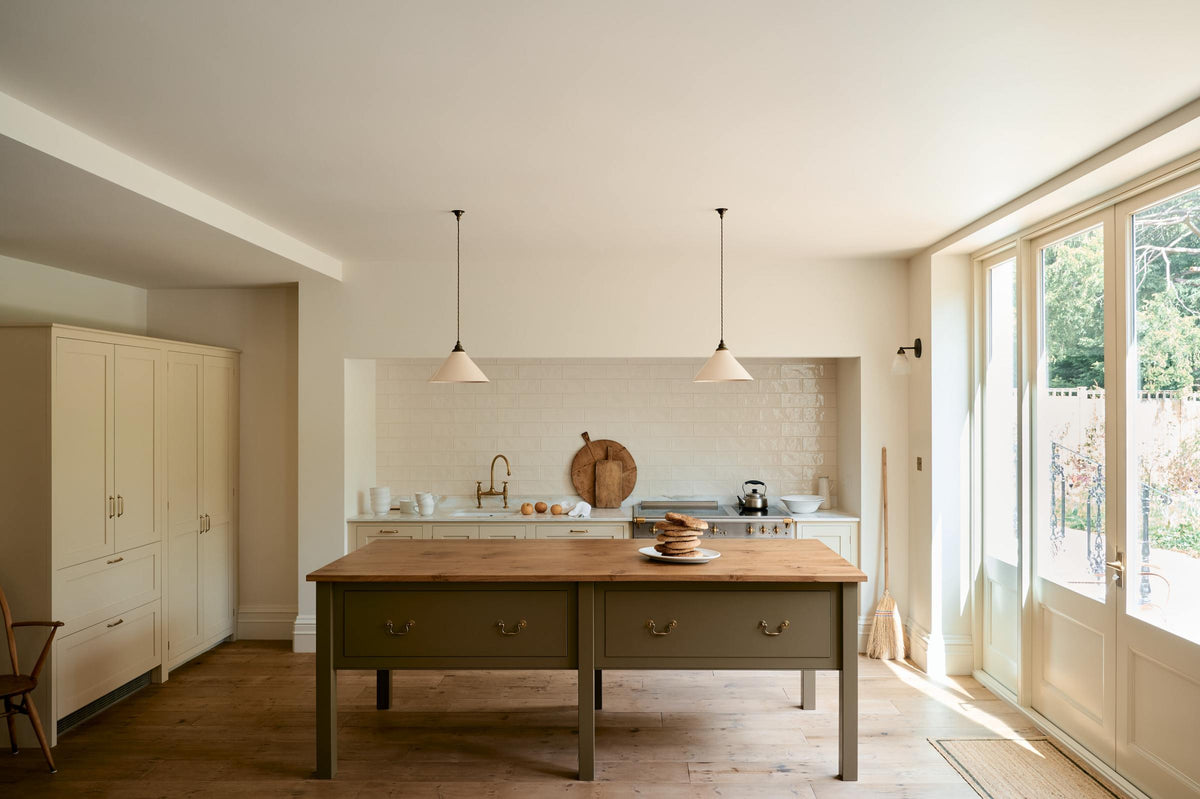 A light, clean, and simple shaker kitchen features a sink and cooker run nestled in a chimney breast, cream porcelain splashback tiles, wooden floorboards, and white walls. Cream cabinets complement an olive green Georgian-style prep table with deep drawers and a rustic oak worktop at the room’s centre, illuminated by two elegant creamware pendant lights.