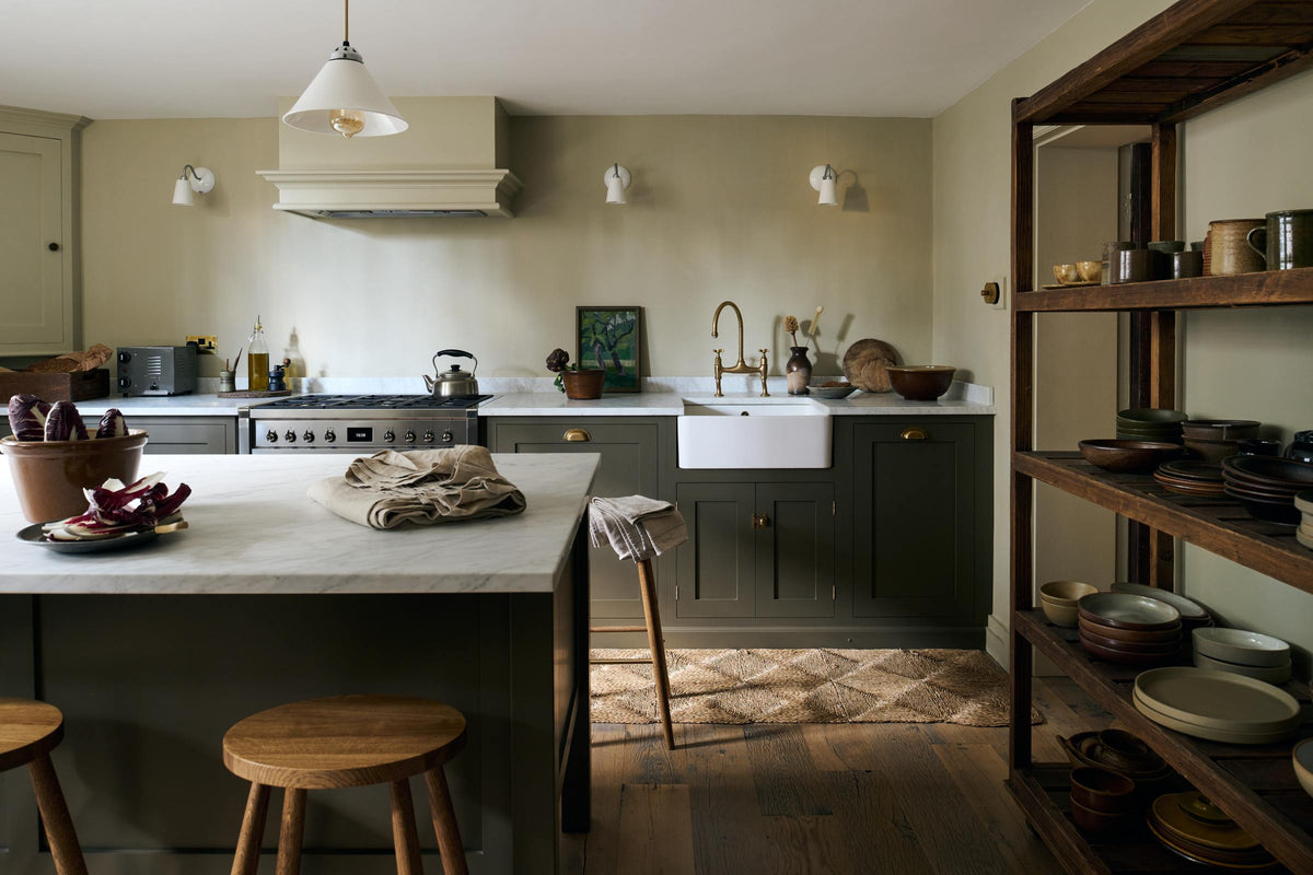 An olive green Shaker kitchen featuring a sink and cooker run, a freestanding prep table with a Carrara marble worktop, and Helen Stools gathered around the breakfast nook. A large wooden shelf displays a thoughtful selection of muted ceramics and mugs. The space embraces a simple theme rooted in raw, natural materials—an ode to Mother Nature—highlighted by a small oil painting of trees placed beside the single Belfast sink with an aged brass Ionian tap.
