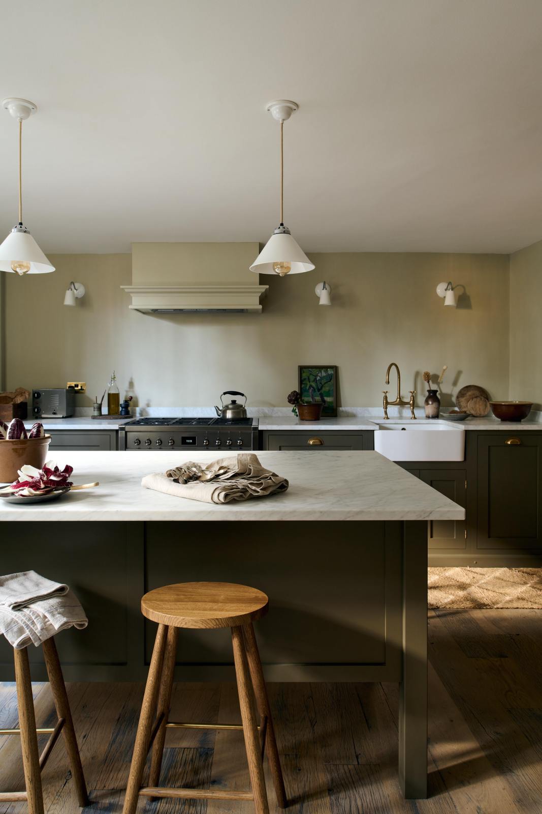An olive green Shaker kitchen featuring a sink and cooker run, a freestanding prep table with a Carrara marble worktop, and Helen Stools gathered around the breakfast nook. A large wooden shelf displays a thoughtful selection of muted ceramics and mugs. The space embraces a simple theme rooted in raw, natural materials—an ode to Mother Nature—highlighted by a small oil painting of trees placed beside the single Belfast sink with an aged brass Ionian tap.