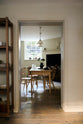 Looking through the archway in the kitchen, beside a large wooden shelf displaying a thoughtful selection of muted ceramics and mugs, the dining room comes into view with a pale wooden dining table and matching chairs.